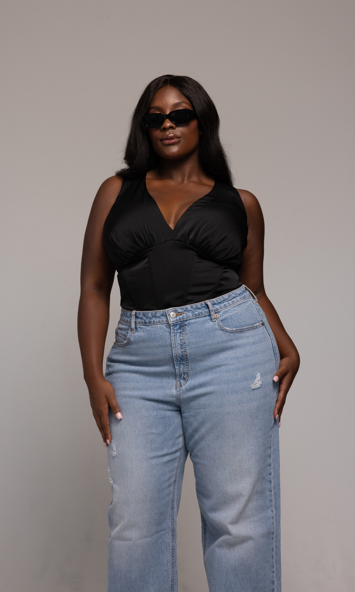A person wearing sunglasses, the GIA/irl Briana Top—a black sleeveless piece with a deep V-neckline—and light blue distressed jeans stands confidently against a plain background. They are posing with a hand on their hip and the other hanging naturally by their side.