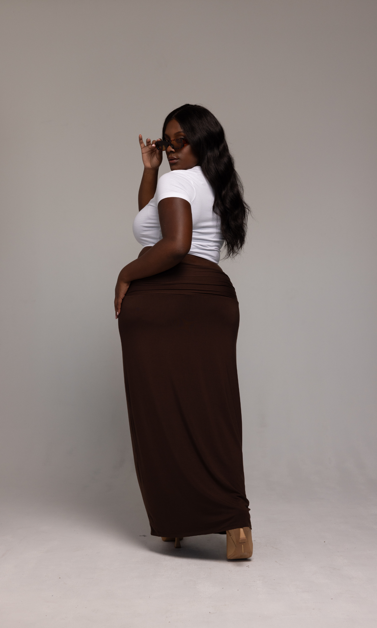 A woman poses confidently in a white top and the Valentina Skirt by GIA/irl, with her back to the camera and head turned slightly. She is wearing glasses and has long, wavy hair. The background is a plain, light grey studio setting.