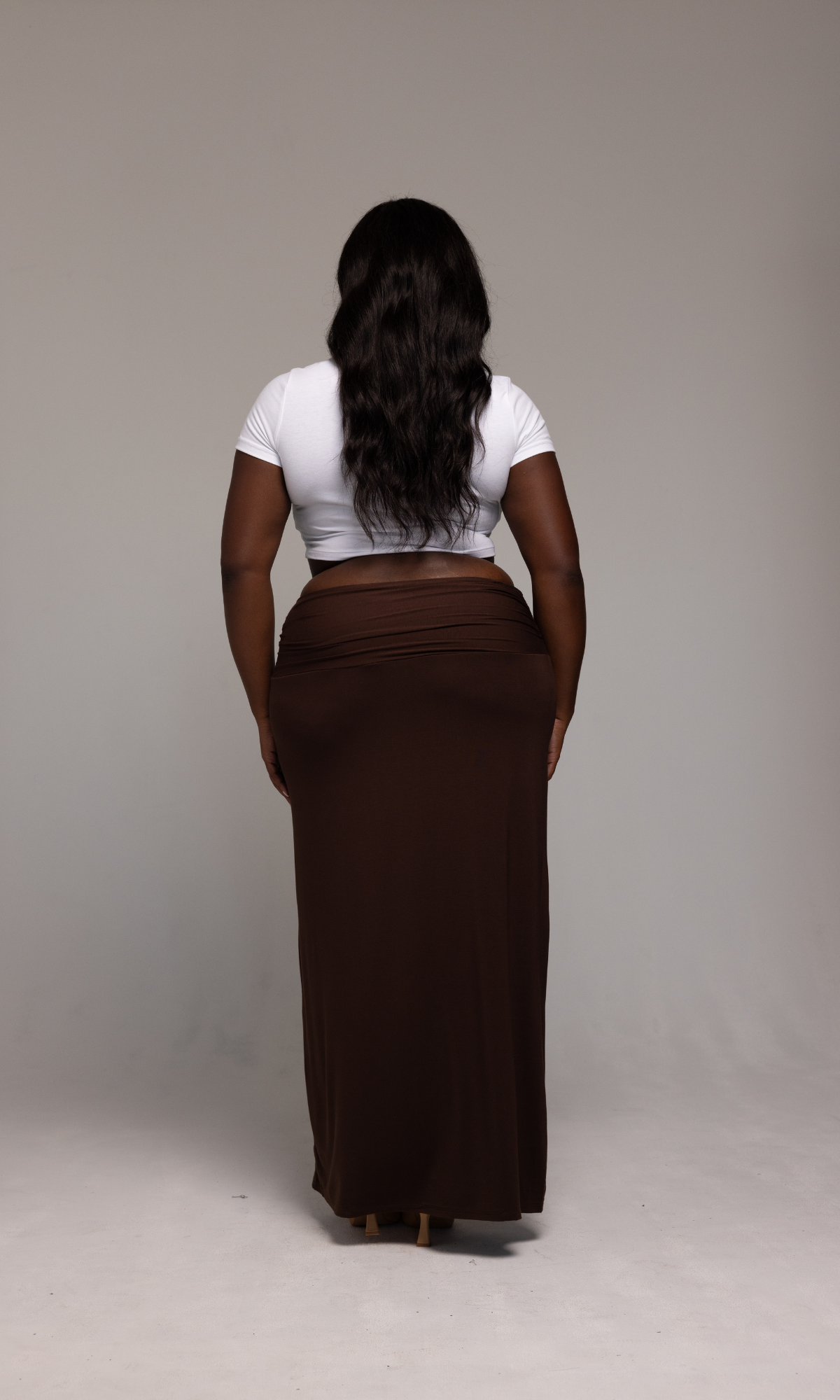 A woman with long, wavy hair stands facing away from the camera. She is wearing a white crop top and the Valentina Skirt by GIA/irl, a long, brown skirt. The background is a plain, neutral color.