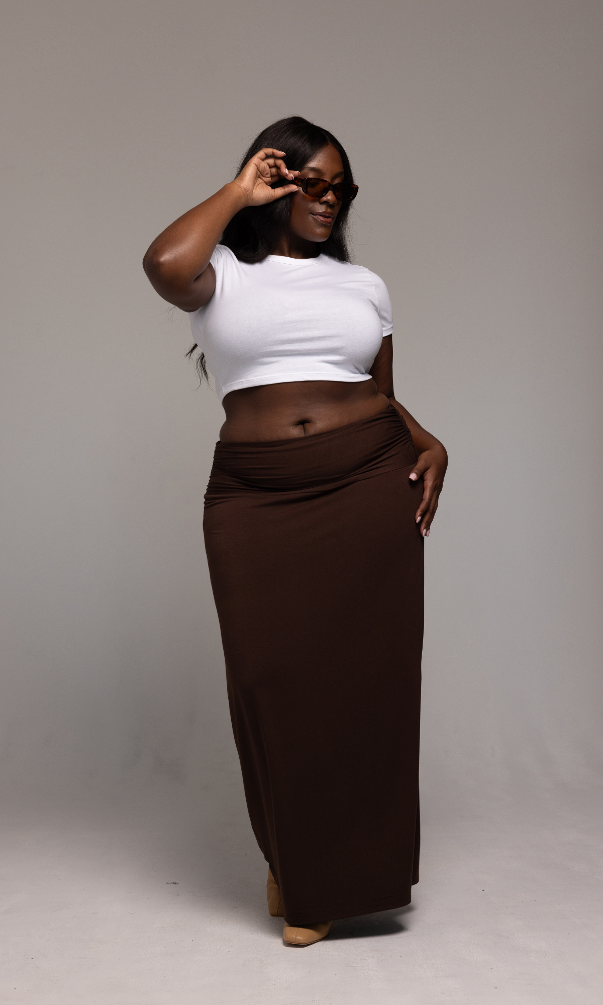 A woman with long hair is posing confidently, wearing sunglasses, a white crop top, and the Valentina Skirt from GIA/irl. She is standing against a plain background and holding the sunglasses with one hand.