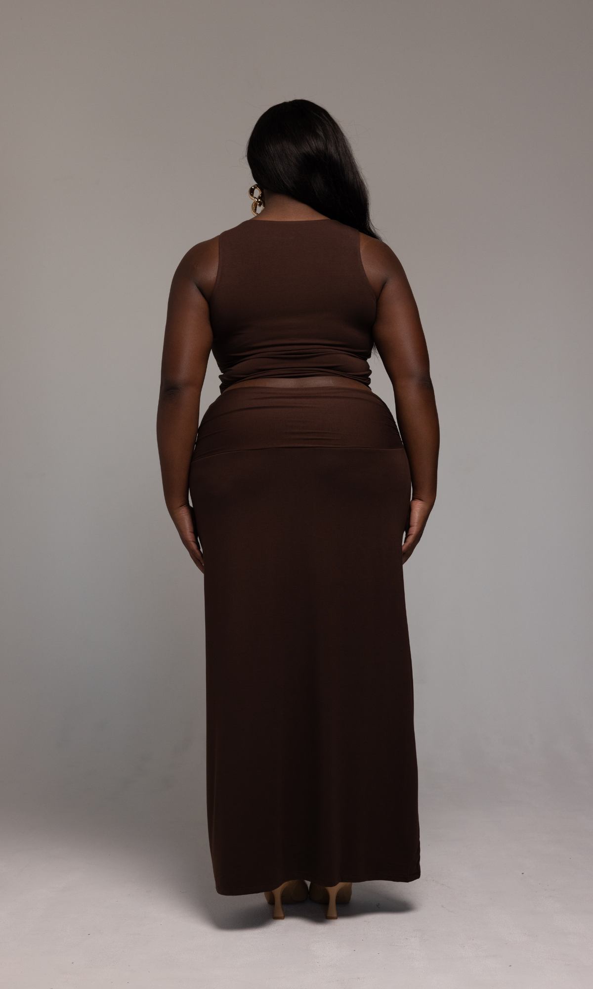 A person stands facing away from the camera, wearing a form-fitting Valentina Top from GIA/irl, paired with a long, sleeveless brown skirt that extends to the floor. They have long, dark hair cascading over their shoulder and are standing against a neutral gray background.