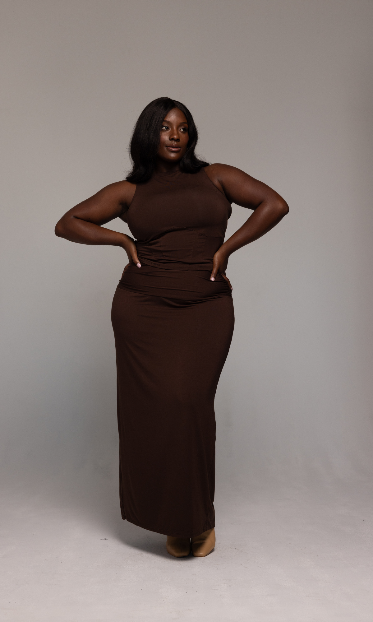A person stands confidently against a neutral background, showcasing the Valentina Top by GIA/irl styled with a sleeveless, form-fitting brown maxi dress and beige shoes. They have long, dark hair and are posing with one hand on their hip and the other slightly behind them.