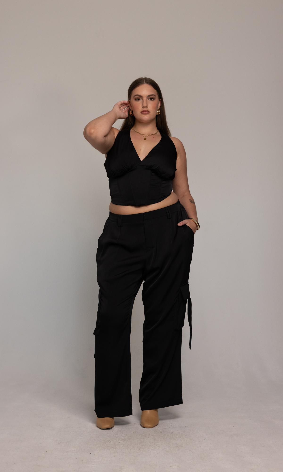 A woman stands confidently against a plain background, dressed in the GIA/irl Briana Pant, paired with a black V-neck crop top. She has straight hair and accessorizes with gold jewelry, including a necklace and bracelets. Striking a pose with one hand on her waist and the other lightly touching her head, she completes her look with beige shoes.