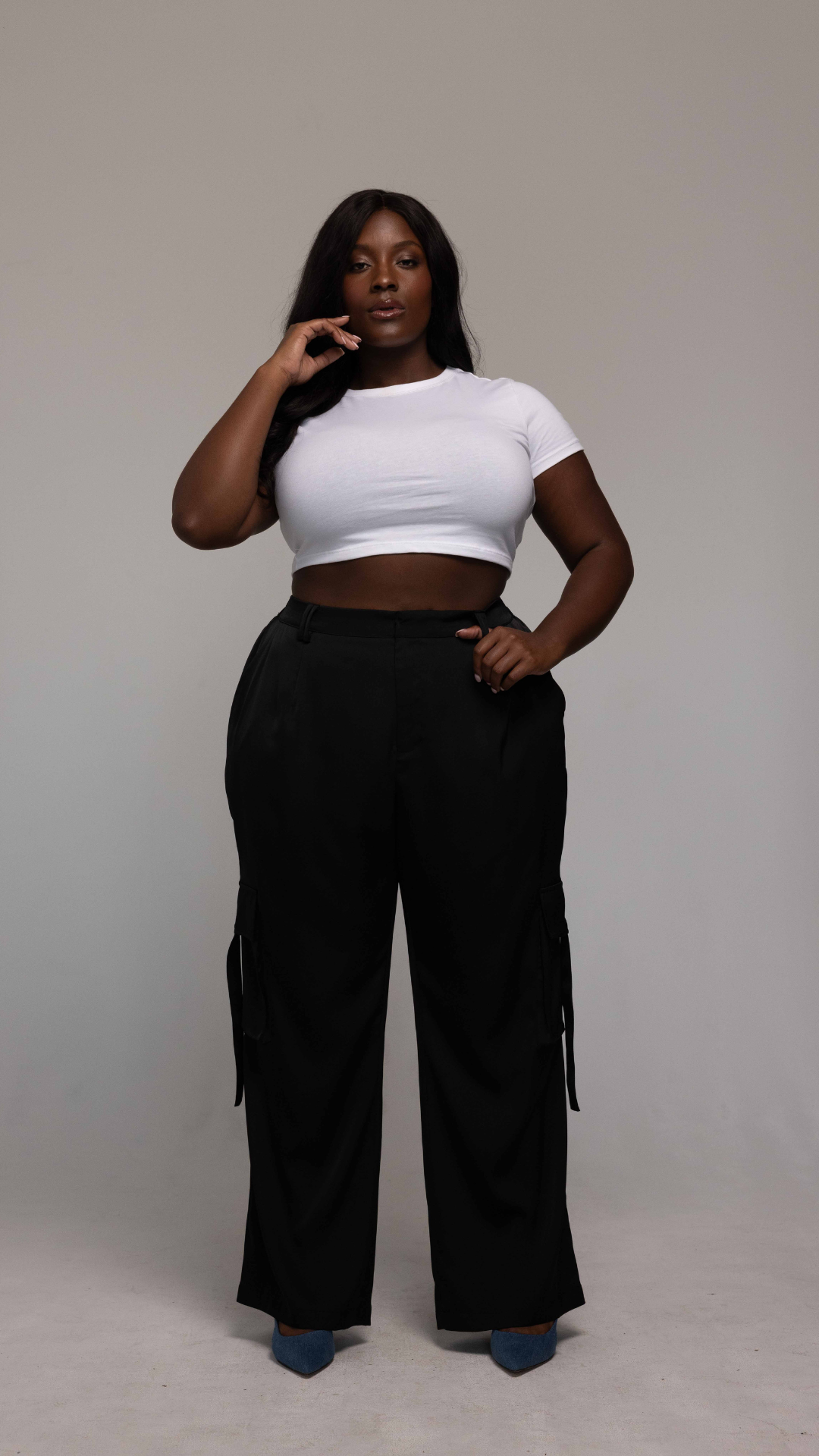 A person with long, dark hair stands confidently against a plain backdrop. They are wearing a fitted white crop top and the high-waisted black Briana Pant by GIA/irl, paired with blue shoes. Their left hand rests on their hip, and their right hand touches their neck.