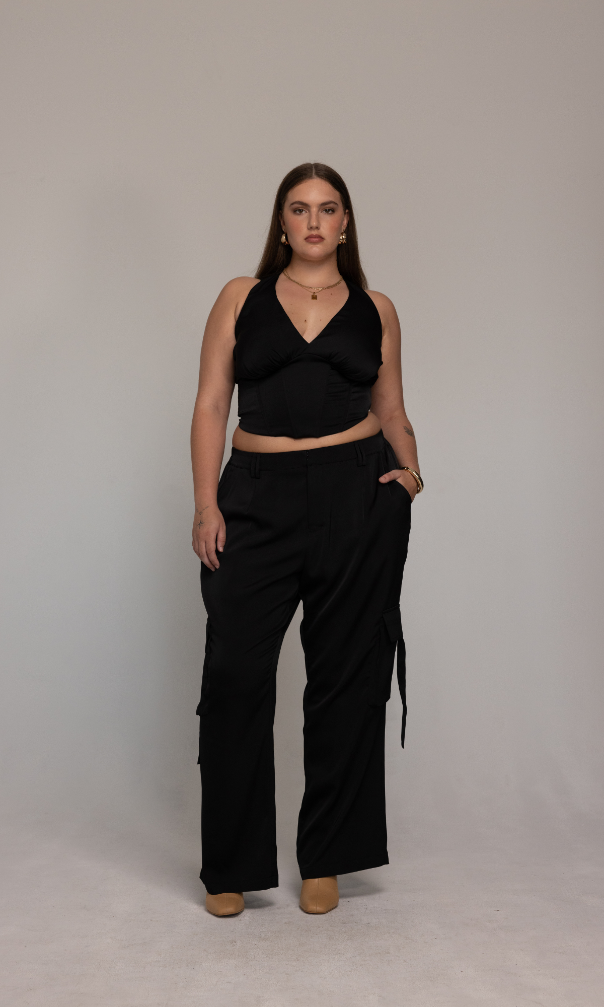 A woman with long brown hair stands against a plain gray background. She is wearing the Briana Top, a black sleeveless V-neck crop top by GIA/irl, and matching black high-waisted pants with pockets. She has beige shoes and accessorizes with a necklace and a watch. She gazes straight at the camera.