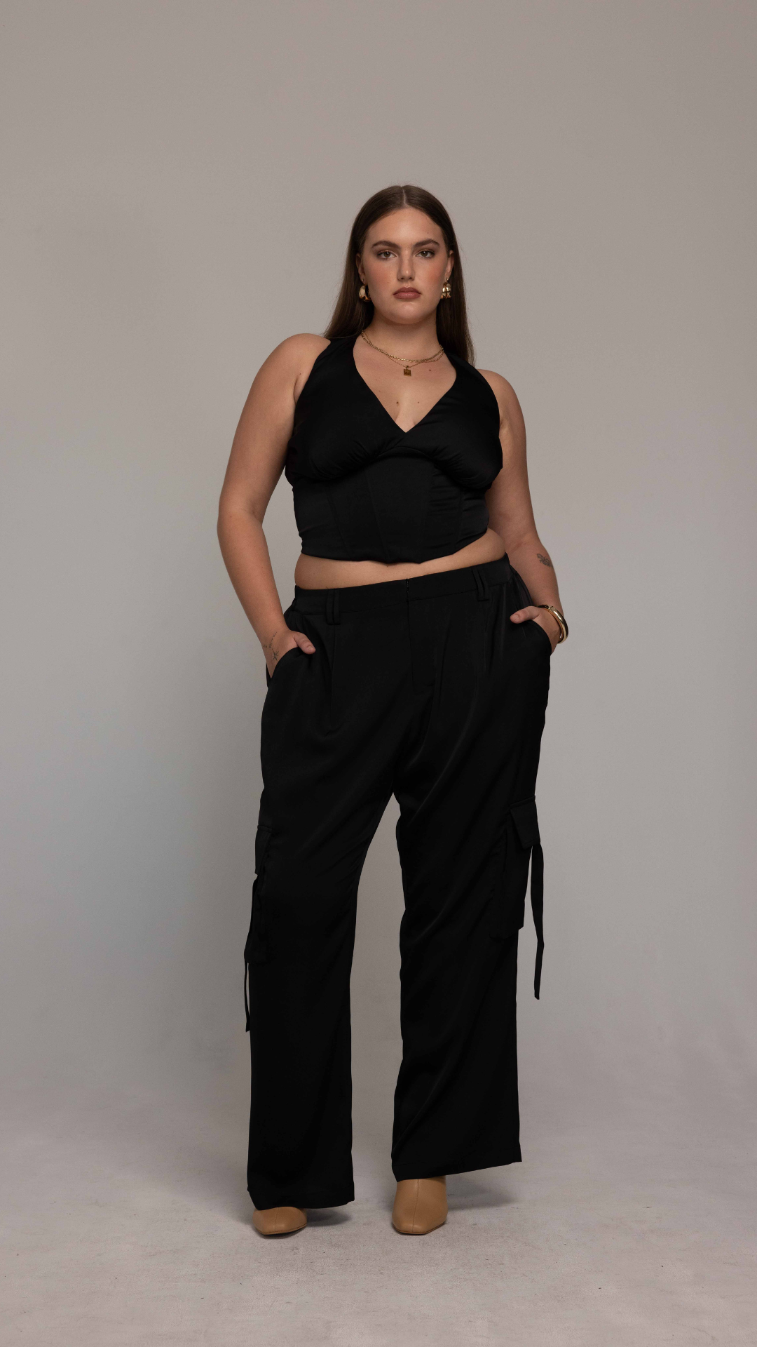 A woman stands confidently against a plain backdrop. She is wearing a black sleeveless crop top and the Briana Pant by GIA/irl, which are high-waisted black cargo pants with pockets. She has long hair, gold earrings, a necklace, a bracelet, and is also wearing tan shoes. Her hands rest comfortably in her pockets.