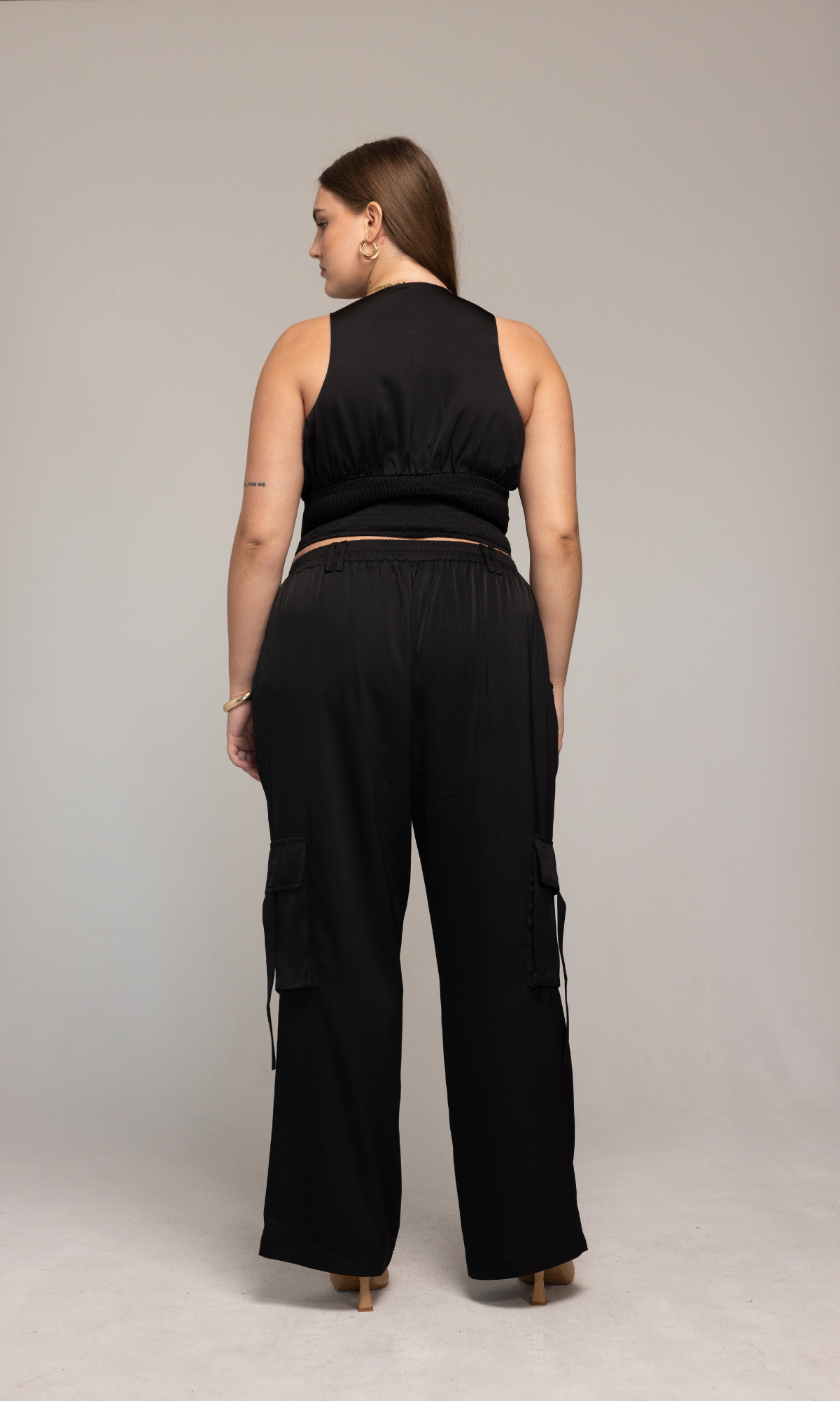 A woman is standing with her back to the camera, wearing the sleeveless black Briana Top by GIA/irl and black cargo pants. The outfit features a cinched waist. Her hair is long and brown, and she has on small hoop earrings. The background is plain and light-colored.