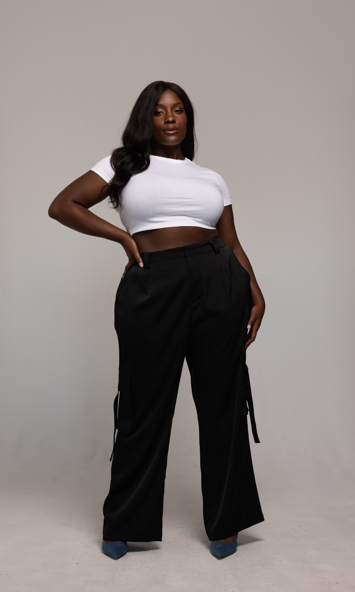 A woman stands confidently against a plain background, wearing a fitted white crop top and the Briana Pant from GIA/irl. The high-waisted black pants feature side pockets and ties. She has long, wavy hair and is posing with one hand on her hip, complementing her outfit with blue heels.