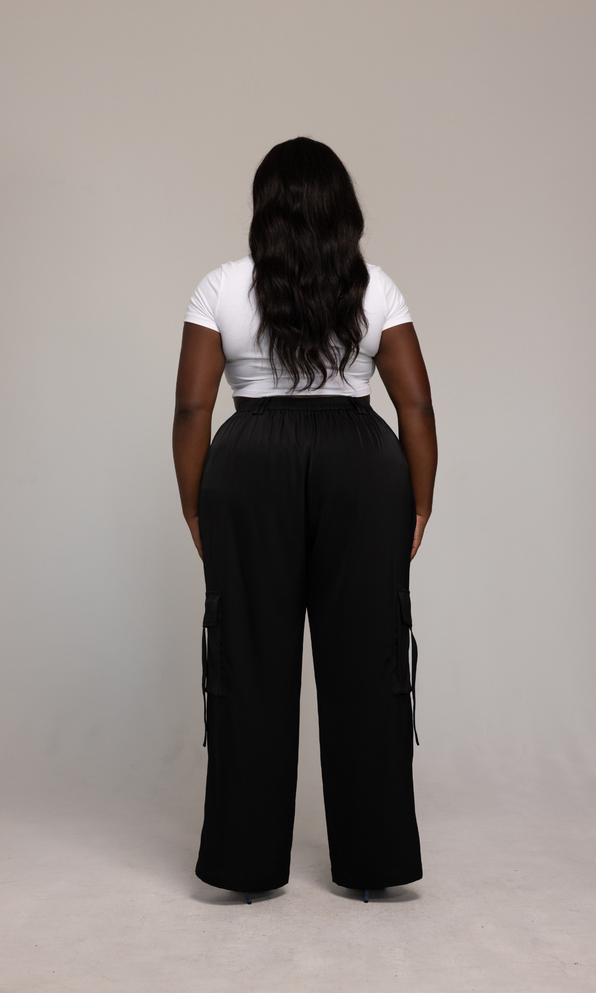 A person with long, wavy hair stands facing away from the camera. They are wearing a white short-sleeve top and the Briana Pant by GIA/irl, which features black high-waisted, wide-leg pants with cargo pockets. The background is plain and light-colored.