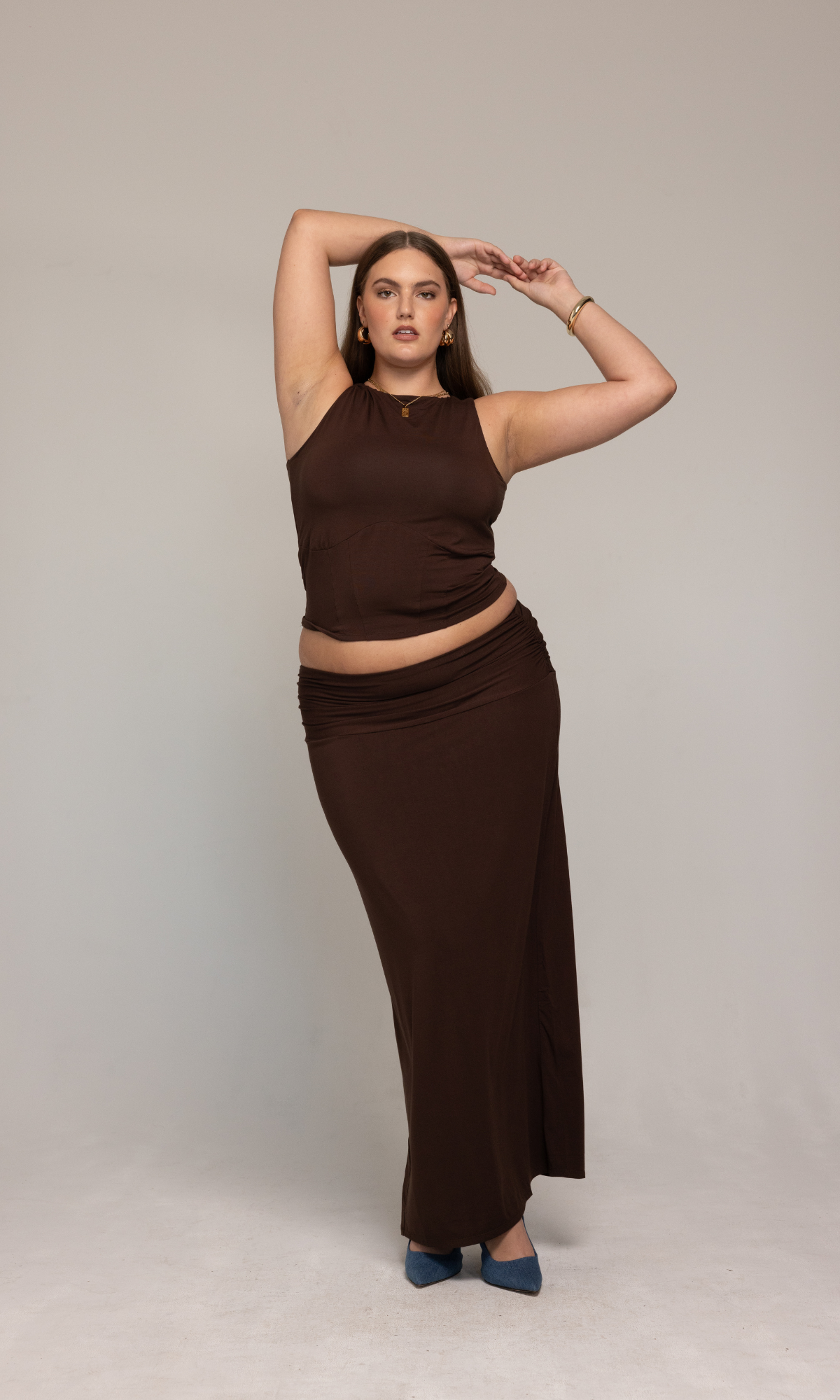 A person poses confidently against a neutral background, wearing a sleeveless brown top paired with the Valentina Skirt by GIA/irl. They have one arm raised above their head and are sporting blue heels. Their makeup is polished, complemented by hoop earrings and bracelets.
