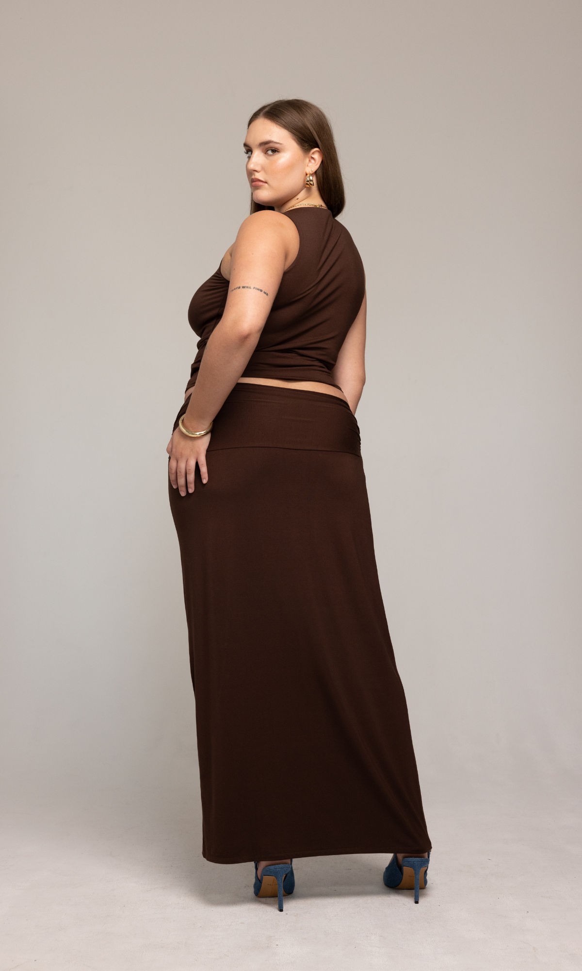 A woman with long hair stands in a studio setting, facing away from the camera. She is wearing the sleeveless Valentina Top from GIA/irl and a matching long brown skirt, paired with teal high heels. She has small tattoos on her arms and is accessorized with gold hoop earrings.