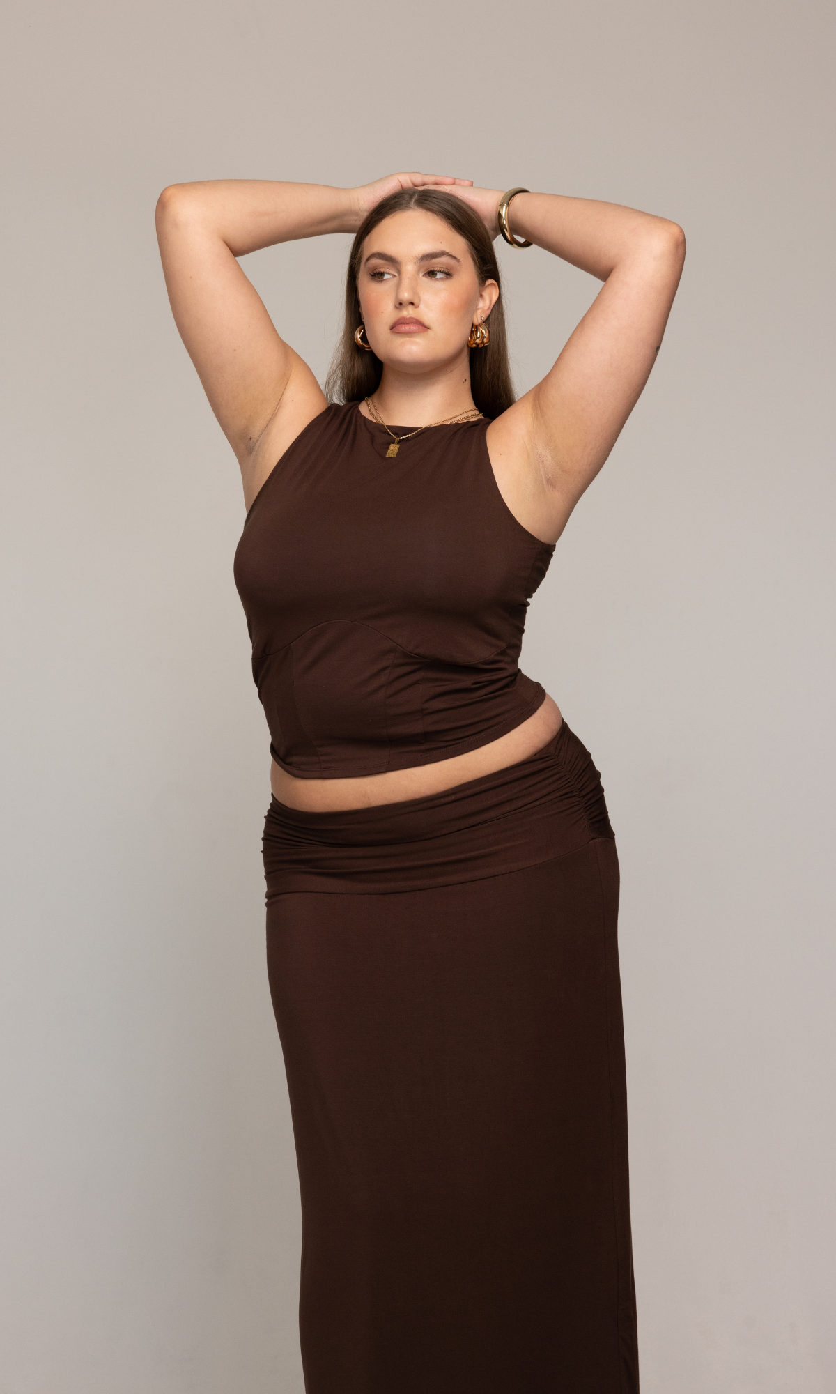 A person stands confidently against a neutral background, wearing the Valentina Top from GIA/irl. They have long, straight hair and are accessorized with gold hoop earrings, a necklace, and a bracelet. One arm rests on their head while the other hangs by their side.
