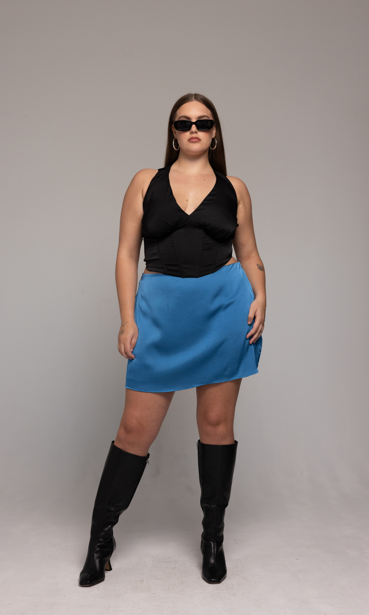 A person with long brown hair is wearing dark sunglasses, the Briana Top from GIA/irl, a blue skirt, and black knee-high boots. They are standing against a plain light gray background.