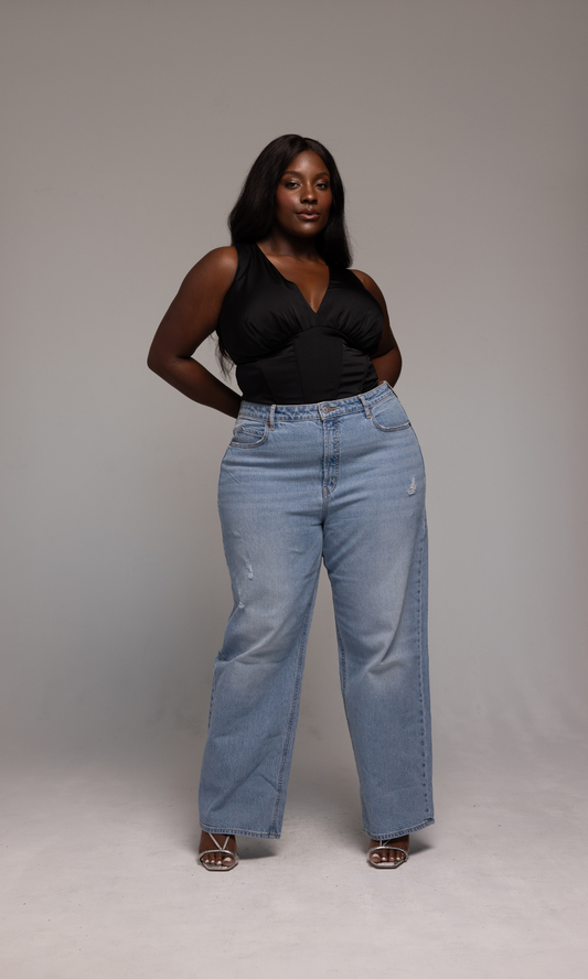 A woman with long black hair is standing confidently against a gray background. She is wearing the Briana Top from GIA/irl, which is a sleeveless black V-neck top, paired with light blue high-waisted jeans and white open-toe heels. Her hands are resting on her hips.