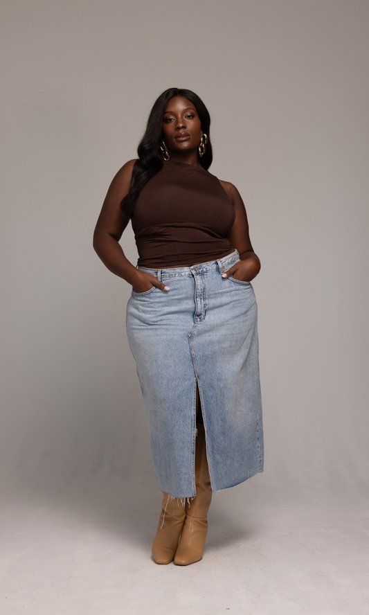 A person stands confidently against a plain background wearing the Valentina Top from GIA/irl, paired with a light blue denim skirt featuring a front slit and tan boots. They have long, dark hair and are accessorized with large hoop earrings. Their hands rest casually in their skirt pockets.
