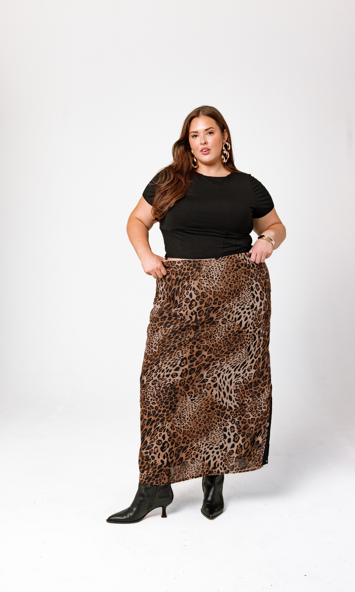 A woman wears a black short-sleeve top, the Bella Leopard Maxi Skirt by GIA/irl, and black ankle boots. She stands in front of a light gray background, posing with her hands on the waistband of her skirt. She has long hair and large hoop earrings.