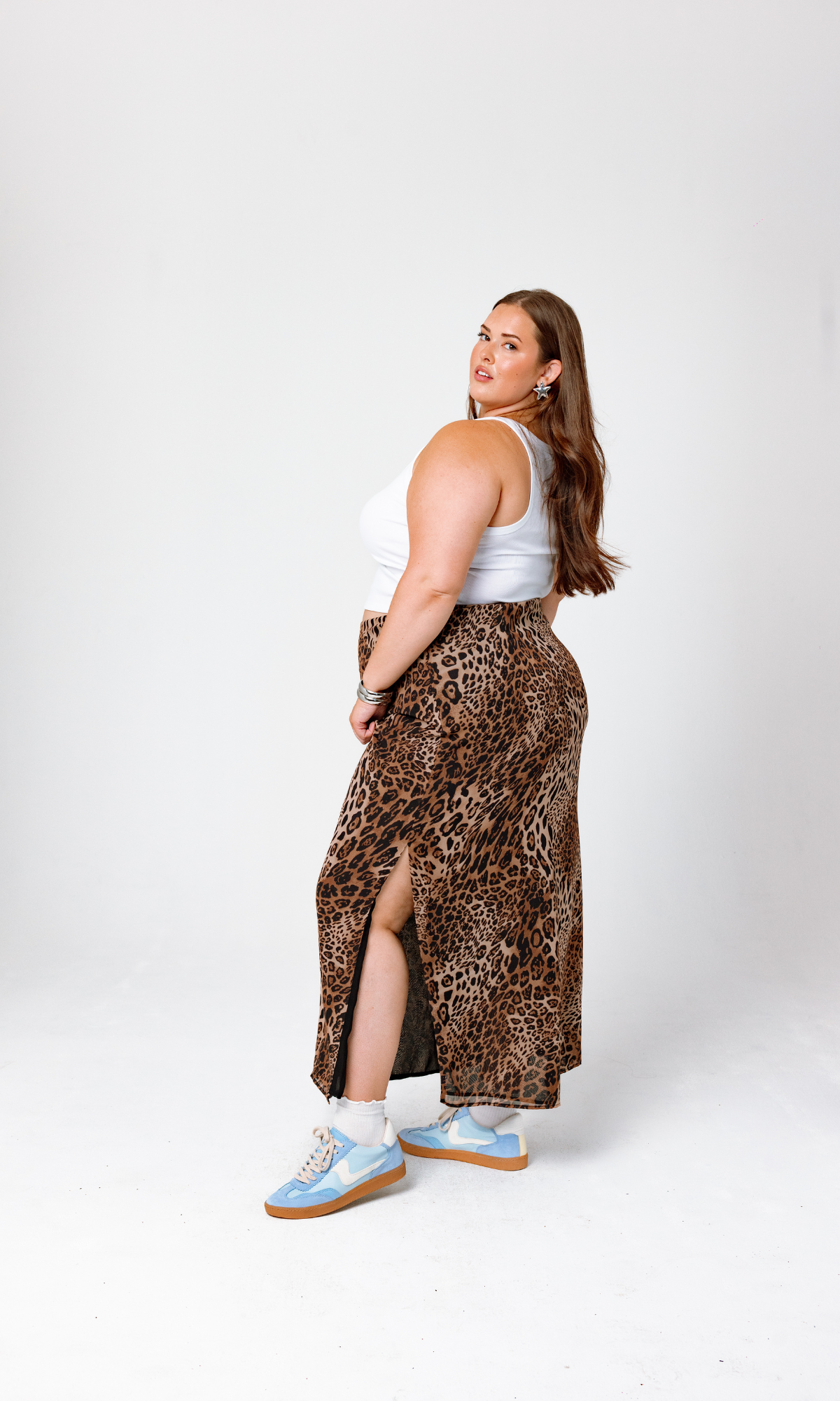 A woman with long brown hair poses against a white background, dressed in a white sleeveless top, the Bella Leopard Maxi Skirt from GIA/irl featuring a side slit, and light blue sneakers. She glances over her shoulder at the camera.