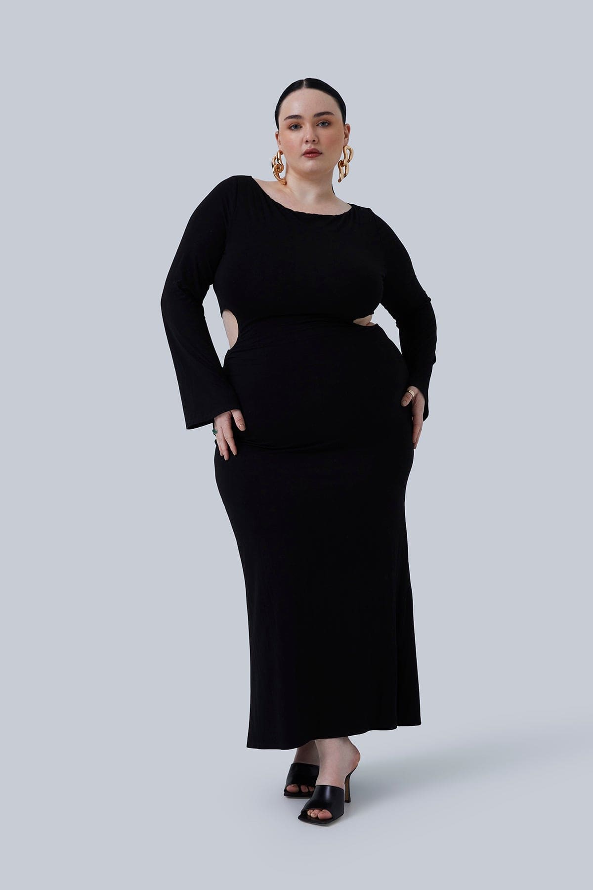 Gabrielle Maxi size 2X in black. Model standing with one leg crossed in front of the other. This long black dress for plus size women has flared sleeves and waist cutouts. Model has slick back hair and big chandelier earrings.