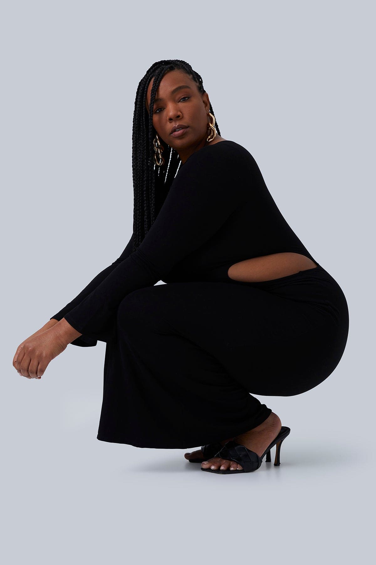 Model squatting from the side in Gabrielle Maxi dress for plus size women. Size oval cutout in this stretchy, double layered fabric. Model has long black braids over her shoulder and is looking straight into the camera.