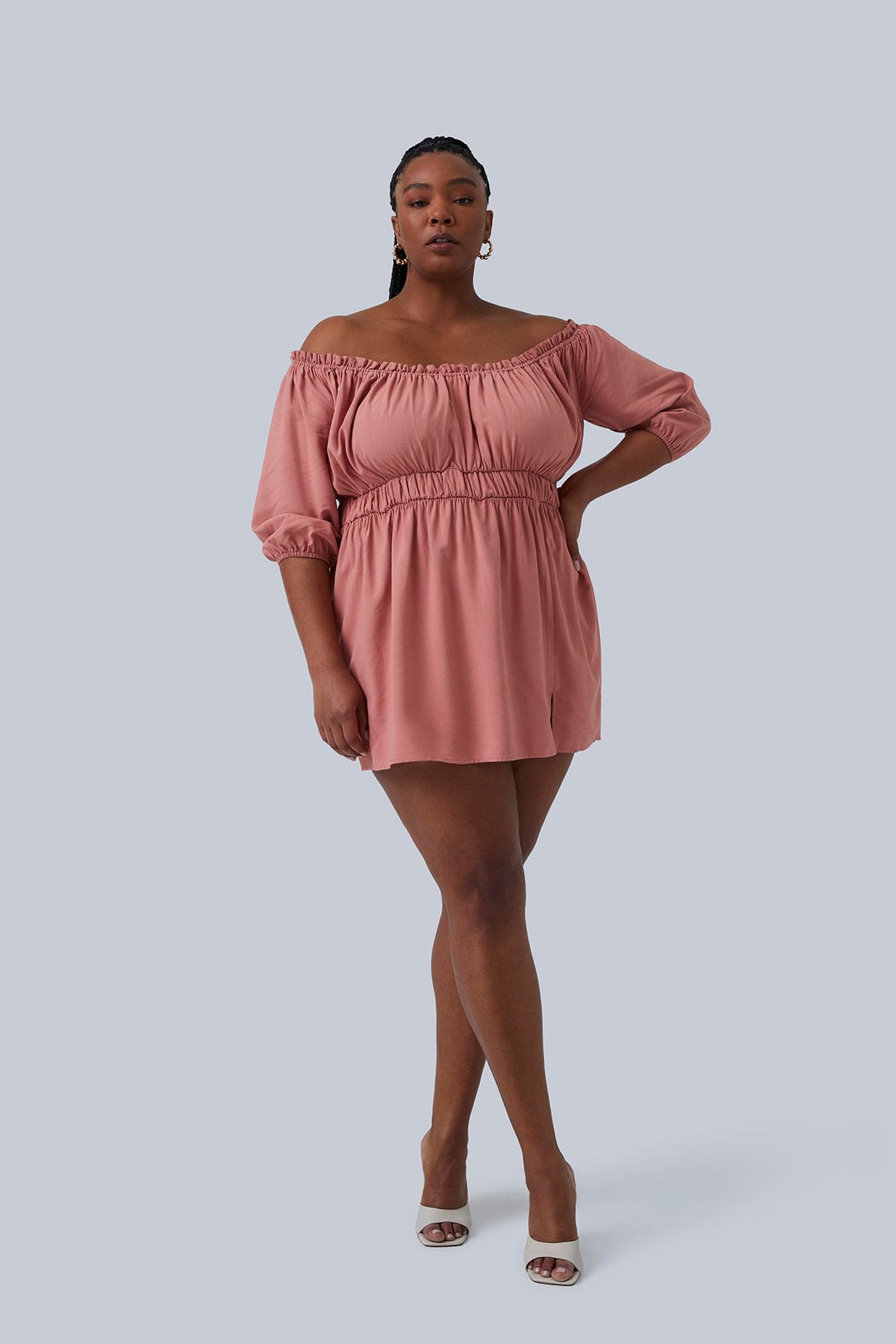 Model standing with hand on hip and legs crossed wearing the Gianna Mini Dress by Gia IRL in a size 1X. Hair is in braids and in a high pony tail showing small gold hoops. Perfect plus size fashion for women. Paired with white peep toe heels.