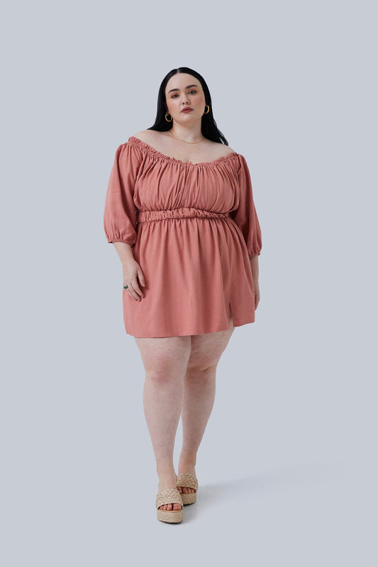 Plus size model is standing wearing the Gianna Mini Dress, her hair is behind her shoulders showing off the off the shoulder detail of this mini dress for plus size women. Color is blush and size is 3X. Shop Gia IRL Plus Size Boutique.