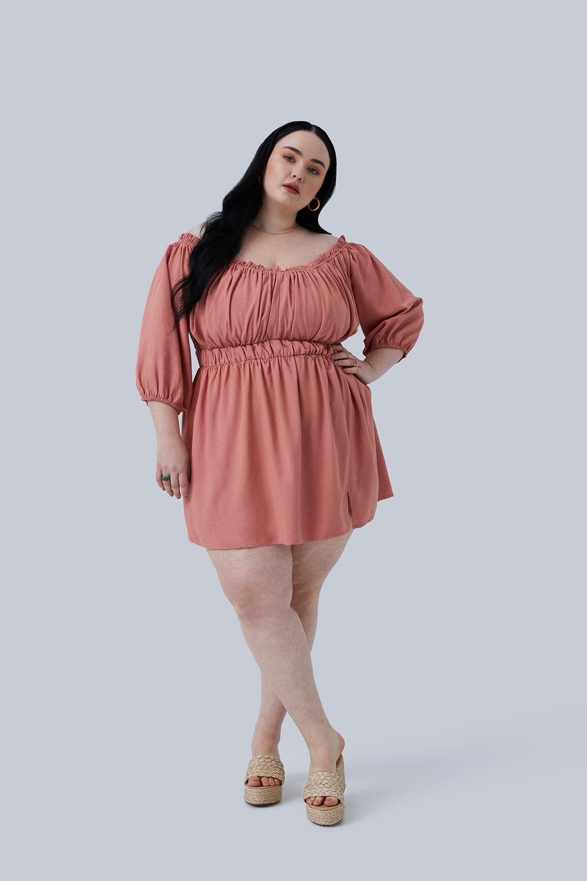 Model standing with one hand on hip and legs crossed wearing the Gianna Mini Dress in blush from Gia IRL Plus Size Boutique in a size 3X. Ruched Off the Shoulder Mini Dress with Ruffle Detail, Blouson Sleeve, and Slit.