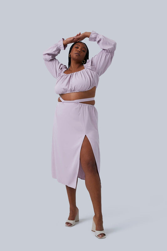 Gia Skirt is a A-Line Midi Skirt with Waist Tie Detail and Thigh High Slit.Model standing with hands over head wearing two piece set for plus size women. The Gia Skirt is in lilac size 2X. Set is sold separately and can be worn separately.