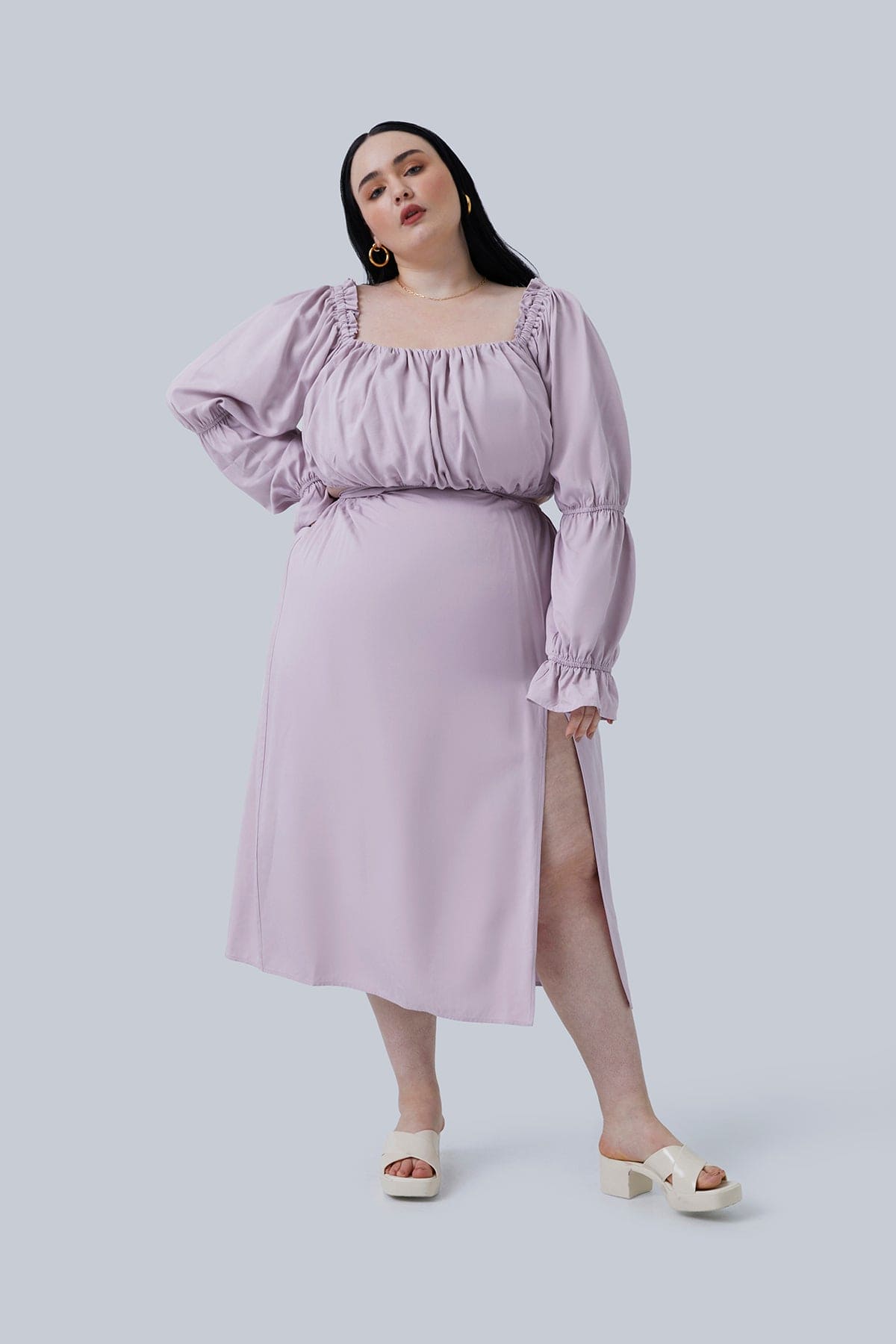 The Gia top in lilac size 3X by Gia IRL Plus Size Boutique. Model is standing with one foot out and one hand on hip, head tilted to the side. You can see the ruched detail and blouson sleeves. The plus size fashion for women you want to shop.